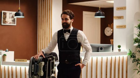 Photo for Cheerful smiling professional bellboy in formal attire with baggage trolley in front of check in reception. Happy hotel employee carrying luggage in resort lobby, reveal jib up shot - Royalty Free Image