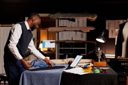 Téléchargez les photos : Suitmaker expert couture ensemble costume d'affaires sur mesure, à la recherche d'inspiration en ligne. Couturier qualifié travaillant dans un atelier professionnel, créant une pièce de vêtement sur mesure de qualité supérieure - en image libre de droit