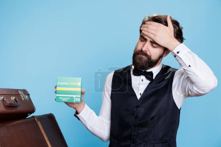 Pförtner zeigt Schachteln mit Schmerzmitteln gegen Kopfschmerzen, während er vor blauem Hintergrund posiert. Männerbellhop hält Medikamentenpackung in der Hand und versucht, Migräne mit Medizinprodukten im Studio zu lindern.