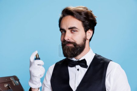 Photo for Rich elegant man spraying perfume, posing with bottle of luxurious cologne and indicating strong masculine scent. Young adult applying fresh fragrance to smell good, hotel concierge employee. - Royalty Free Image