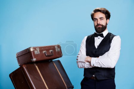 Téléchargez les photos : Travailleur formel de l'hôtel posant avec des bagages, portant un costume formel et des gants blancs sur fond bleu. Homme bellhop debout à côté de pile de sacs et valises de tolley, l'industrie hôtelière. - en image libre de droit