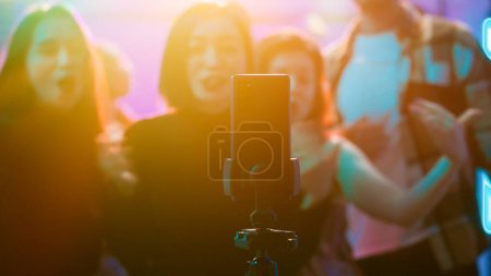 Influencer filming vlog at club party, using smartphone to record video of her and her friends having fun on dance floor. Happy woman enjoying night out clubbing with people, social media.
