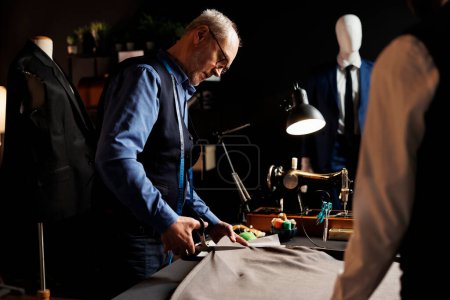 Photo for Precise experienced elderly suitmaker manufacturing suit in tailoring studio workspace, cutting fabric material with scissors. Process of manufacturing upcoming bespoke fashion design collection - Royalty Free Image