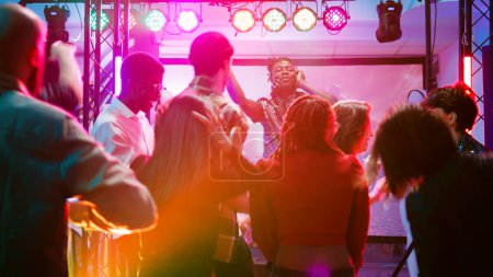 Foto de Grupo multiétnico de personas de fiesta en la pista de baile, disfrutando de la música funky electrónica de DJ estación de mezcla. Adultos alegres divirtiéndose en la discoteca, bailando juntos en la celebración. Disparo de mano. - Imagen libre de derechos