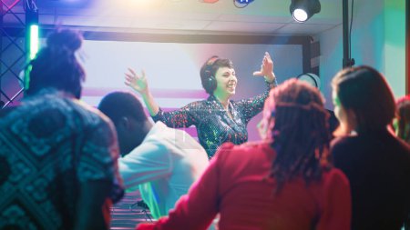 Téléchargez les photos : Jeune DJ mélangeant musique funky à la fête, se sentant joyeux avec des mouvements de danse et des sons électroniques à la station sur scène. Groupe d'amis faisant la fête et dansant ensemble au club, vie nocturne. - en image libre de droit