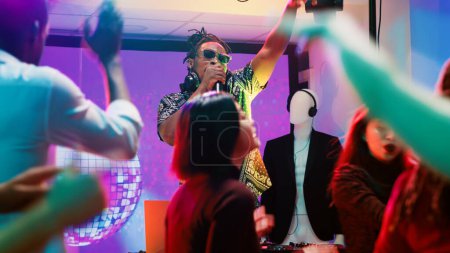 Foto de DJ masculino mezclando música electrónica en la fiesta, divirtiéndose con movimientos de baile y audio moderno en la estación del panel en el escenario discoteca. Gente de fiesta y saltando juntos en el club nocturno, entretenimiento. - Imagen libre de derechos