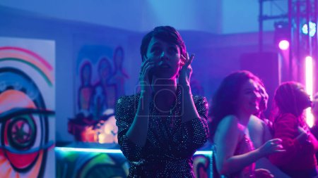 Woman answering phone call at party in the club, trying to have conversation with loud music on dance floor. Young adult using mobile phone remote chat at clubbing event, entertainment.
