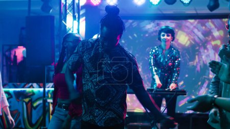 Téléchargez les photos : Groupe d'amis dansant dans le club, profitant d'une performance DJ en direct sur piste de danse colorée. Jeunes gens funky s'amuser à la boîte de nuit avec des lumières disco et de l'alcool, clubbing. Trépied. - en image libre de droit