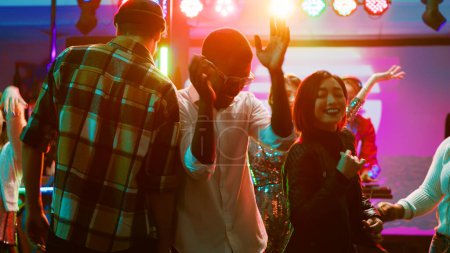 Photo for Diverse friends dancing on live music, feeling cheerful on dance floor at nightclub. Young people having fun with performance at discotheque, doing funky dance moves under stage lights. Tripod shot. - Royalty Free Image