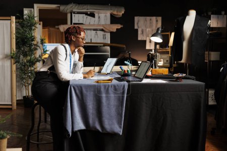 Photo for African american seamstress in atelier shop workspace surrounded by sewing instruments looking online for design inspirations, inspecting sketch drawings of upcoming sartorial collection - Royalty Free Image