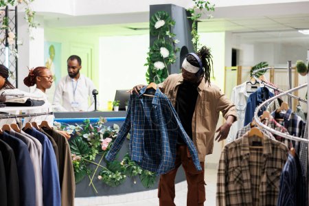Photo for Shopper enjoying music in headphones while browsing for trendy clothes in mall. Showroom customer in earphones holding casual plaid shirt on hanger to try on in fitting room - Royalty Free Image