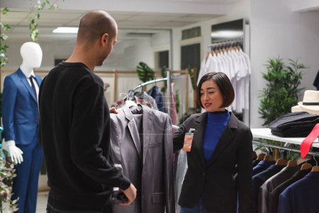 Photo for Fashion boutique arab customer asking asian woman assistant for opinion while choosing formal jacket. Clothing store seller and buyer holding apparel on hanger and discussing style - Royalty Free Image