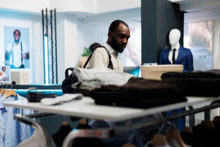 Téléchargez les photos : Homme afro-américain naviguant à travers rack de vêtements décontractés dans le département de la mode de centre commercial. Client choisissant la tenue et vérifiant les vêtements sur les cintres lors du magasinage en boutique - en image libre de droit
