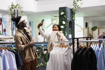 Téléchargez les photos : Femme afro-américaine client prendre selfie sur smartphone tout en choisissant des vêtements dans le centre commercial. Boutique de mode clients essayant l'usure formelle et de faire des photos sur téléphone mobile - en image libre de droit