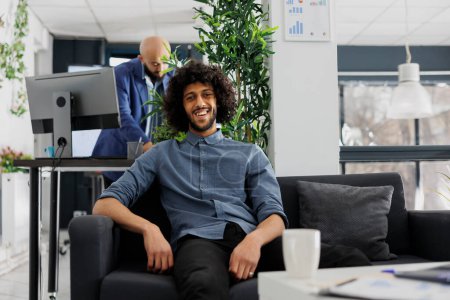 Smiling successful arab start up company entrepreneur in business office portrait. Young handsome executive manager looking at camera, sitting on couch in green coworking space