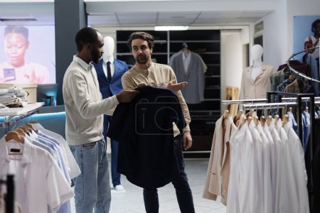 Photo for African american man holding shirt on hanger and asking shopping center worker about size options. Boutique worker pointing at new collection apparel rack, helping customer to choose outfit - Royalty Free Image