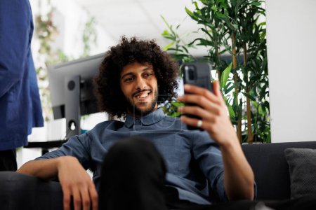 Téléchargez les photos : Entrepreneur arabe réussi parler sur vidéocall smartphone avec un ami pendant la pause de travail dans le bureau d'affaires. Happy relaxed directeur exécutif des employés de l'entreprise bavarder sur téléphone mobile - en image libre de droit