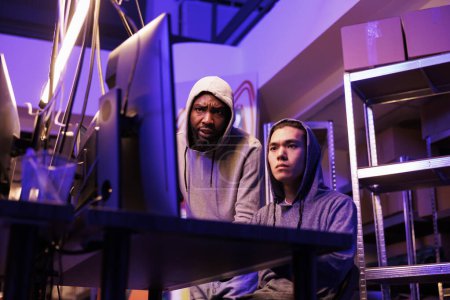 Two concentrated hackers collaborating to crack complex password and break into network system. Young diverse men in hoods working on computer together and coding virus malware