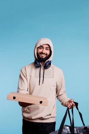 Foto de Servicio de entrega de comida sonriente mensajero árabe con caja de pizza y retrato de mochila. Joven alegre en la capucha dando comida rápida para llevar orden y mirando a la cámara con expresión despreocupada - Imagen libre de derechos