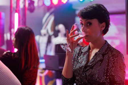 Photo for Young woman drinking alcohol and looking at camera while attending party in nightclub. Cheerful girl holding beverage glass while having fun and enjoying nightlife on dancefloor in club - Royalty Free Image