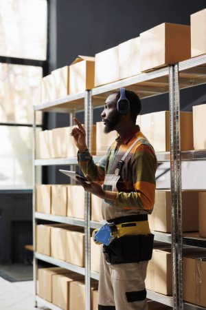 Photo for Delivery service manager in headphones searching parcel and counting cardboard boxes on shelf using inventory app. Shipment operator standing in warehouse aisle, listening to music and holding tablet - Royalty Free Image