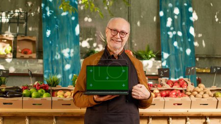Elderly local farmer holding laptop with isolated greenscreen outdoors at local food fair, sunny day. Senior man showing chroma key display with blank mockup copyspce on screen, wearing pron.