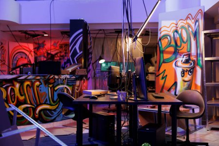 Internet criminals abandoned warehouse with graffiti at night time. Computers on desks and equipment for hacking and malware coding in hackers dark hideout room with neon light