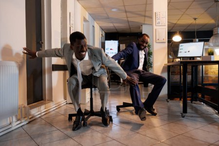 Photo for Playful businessmen riding desk chair during sports race competition during break time in modern startup office. Cheerful african american employees having fun, enjoying doing moving activity - Royalty Free Image