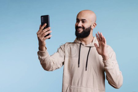 Téléchargez les photos : Homme arabe souriant saluant tout en parlant en ligne à l'aide de l'application de vidéocall smartphone. Personne amicale ayant la communication à distance, saluant avec la main et parlant avec la caméra avant de téléphone portable - en image libre de droit