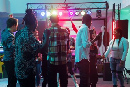 Young people enjoying concert on dancefloor with spotlights during live music performance in nightclub. People crowd clubbing and dancing while attending party at nighttime