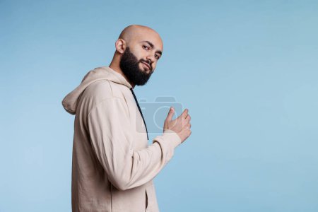 Smiling arab man inviting with hand and looking at camera with relaxed facial expression. Young bald bearded person asking to come over with arm gesture, posing for studio portrait