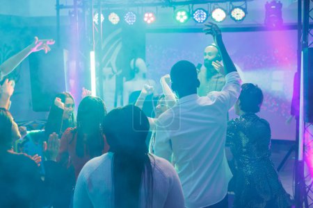 Photo for People jumping and raising hands while partying at dj performance in dark nightclub. Crowd clubbing, dancing and having fun on dancefloor at electronic music concert in club - Royalty Free Image
