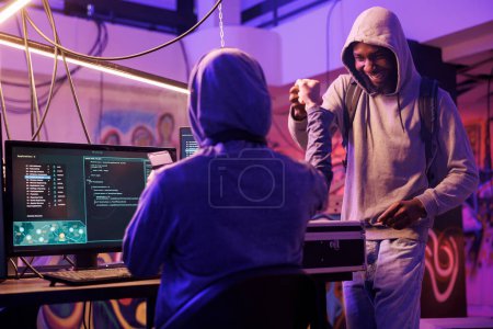 Photo for Cheerful african american hacker giving fist bump to crime partner while working together in abandoned warehouse. Smiling men in hoods sharing friendly gesture after successful database breaking - Royalty Free Image