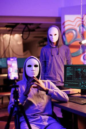 Scammers in anonymous masks streaming threat online and demand victim for payment. Hackers with hidden identity filming ransom message on mobile phone in abandoned warehouse