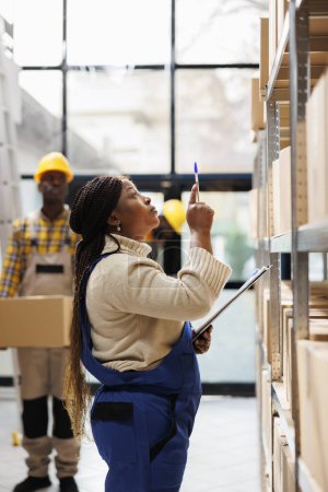 Photo for African american woman warehouse manager looking at parcels and controlling goods stock. Logistics operator coordinating products storage and taking notes near cardboard boxes shelf - Royalty Free Image