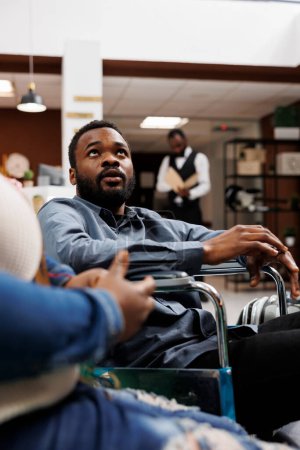 Junger afroamerikanischer Mann im Rollstuhl bei der Ankunft im Luxushotel mit behindertengerechten Einrichtungen, behinderter Mann, der mit seiner Frau unterwegs ist. Barrierefreier Tourismus, Reiseunterkünfte und Behinderungen