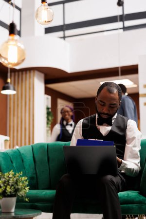 Photo for Computer and hotel industry. Black guy bellboy sitting with laptop in lobby entering data filling online document. African American man concierge making entry in daily luggage movement register - Royalty Free Image