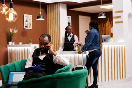 Téléchargez les photos : Jeune homme afro-américain gestionnaire d'hôtel assis dans le hall parler par téléphone et prendre des notes, la gestion des réservations en ligne. noir guy groom tenant smartphone commander taxi pour invité - en image libre de droit
