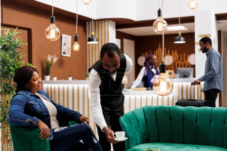 Photo for Happy smiling African American woman tourist relaxing in hotel lobby, looking at waiter serving coffee, feeling satisfied with customer service. Friendly restaurant worker putting cup on table - Royalty Free Image