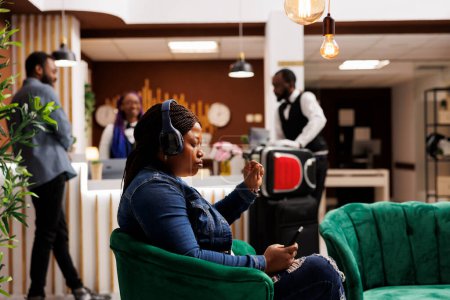 Foto de Una turista afroamericana seria que usa auriculares usando un teléfono inteligente mientras está sentada en el salón del hotel contra la recepción. Invitada femenina enfocada completando el proceso de check-in en el dispositivo móvil - Imagen libre de derechos