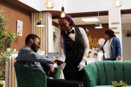 Téléchargez les photos : Un touriste afro-américain souriant tenant une facture par carte de crédit tout en se reposant dans le salon de l'hôtel. Serveuse sympathique tenant pos terminal servant invité dans le hall de l'hôtel - en image libre de droit