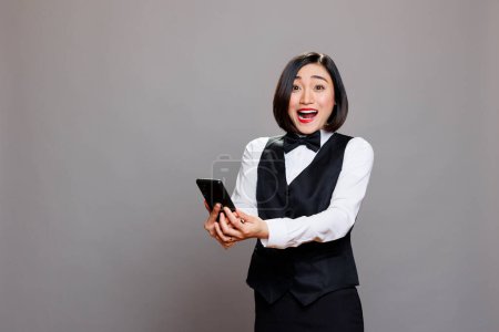 Téléchargez les photos : Excité heureux asiatique femme hôtel réceptionniste en uniforme tenant smartphone et regardant caméra. Surpris jeune serveuse souriante lisant de bonnes nouvelles sur le portrait de téléphone mobile - en image libre de droit