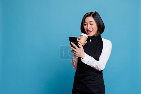 Photo for Cheerful asian waitress laughing while drinking takeaway coffee and scrolling social media on smartphone. Smiling woman receptionist enjoying beverage to go and using mobile phone - Royalty Free Image