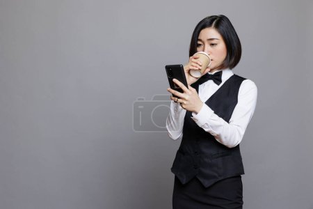 Téléchargez les photos : Jeune femme asiatique portant uniforme de serveuse tapant sur smartphone et profiter de prendre du café. Réceptionniste restaurant utilisant téléphone portable et boisson de tasse en papier - en image libre de droit
