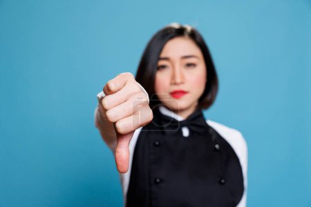 Photo for Young asian woman wearing waitress uniform showcasing disapproval with thumb down gesture portrait. Restaurant receptionist showing service negative feedback concept and looking at camera - Royalty Free Image