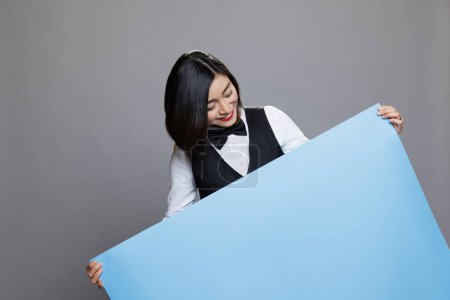 Photo for Smiling asian waitress wearing restaurant uniform holding blue empty paper banner in hands. Catering service young joyful woman employee posing with blank promotion poster - Royalty Free Image