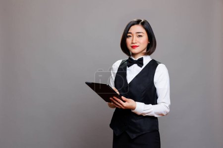 Photo for Smiling confident asian waitress wearing professional uniform, holding digital tablet and looking at camera. Young attractive hospitality industry receptionist using portable gadget portrait - Royalty Free Image
