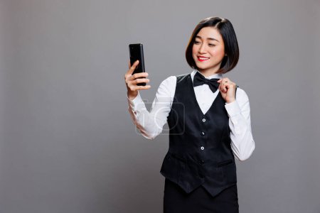 Téléchargez les photos : Souriant asiatique serveuse bavarder en appel vidéo avec clleague en utilisant smartphone. Femme gaie réceptionniste arrangeant uniforme noeud papillon tout en parlant en conférence en ligne sur téléphone mobile - en image libre de droit