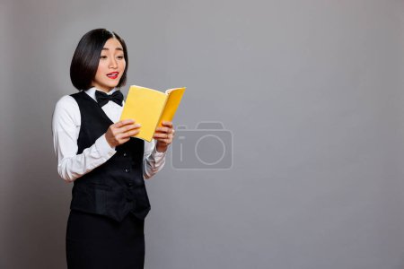 Photo for Surprised young asian waitress reading textbook with yellow cover. Excited reception woman employee wearing black and white uniform holding softcover book, learning literature - Royalty Free Image