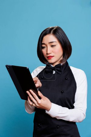 Téléchargez les photos : Restaurant jeune employée asiatique portant uniforme tapant sur tablette numérique. Cafétéria serveuse attrayante répondant au message en ligne du client sur gadget portable en studio - en image libre de droit
